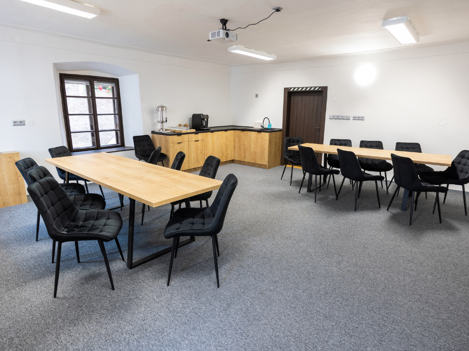 Meeting room with two long wooden tables and black cushioned chairs, kitchenette in back and ceiling projector above.