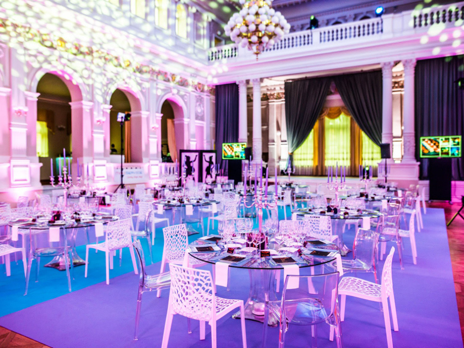 Elegant banquet hall set for a formal reception with round tables, glassware, and white chairs under pink-purple lighting and chandeliers
