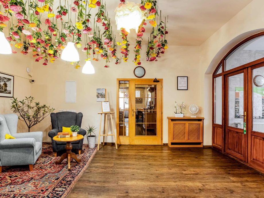 Reception lobby with colorful flower garlands overhead, two armchairs, a wooden table, and a rug; wooden doors and a cabinet in the background.