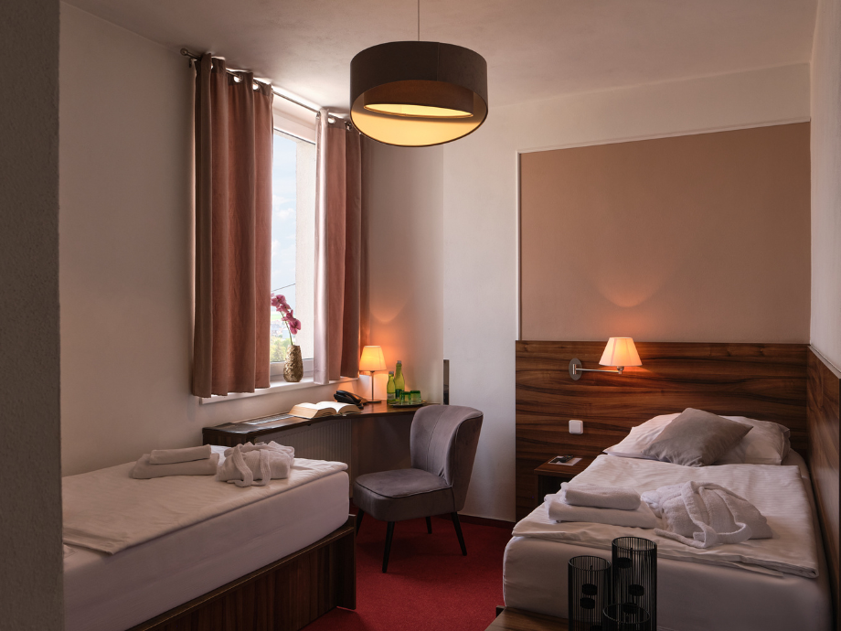 Twin hotel room with two single beds, a desk, chair, and warm lighting from bedside lamps by the window with brown curtains.
