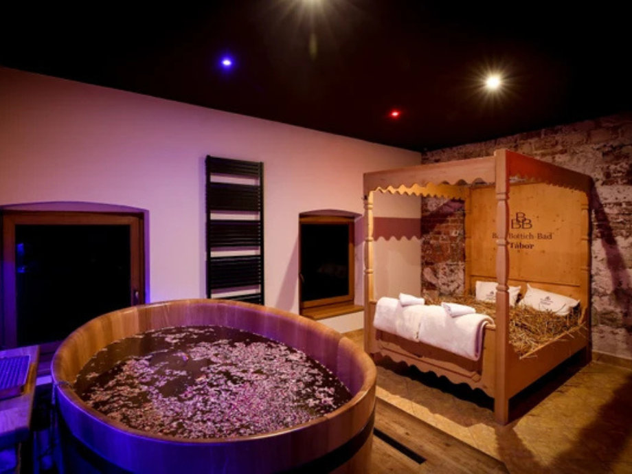 Spa room with a round wooden soaking tub filled with water and petals, next to a four-poster bed covered with towels under warm lighting.