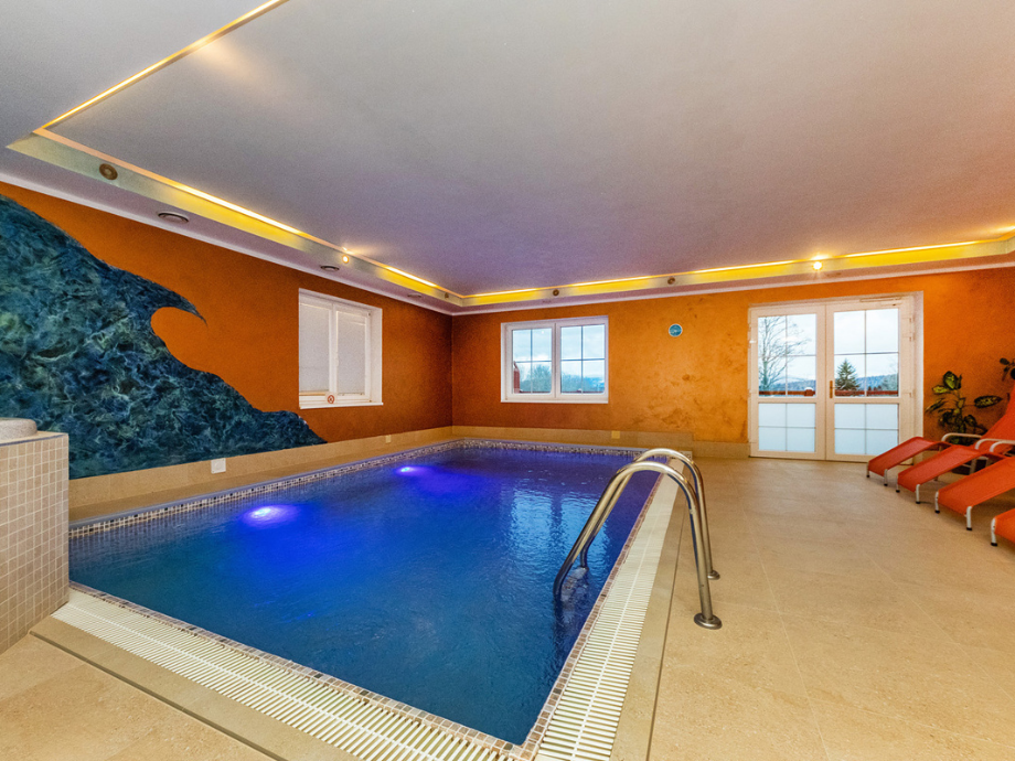 Indoor pool area with blue water, orange walls, and a wave mural on the left; orange lounge chairs line the right side near glass doors.