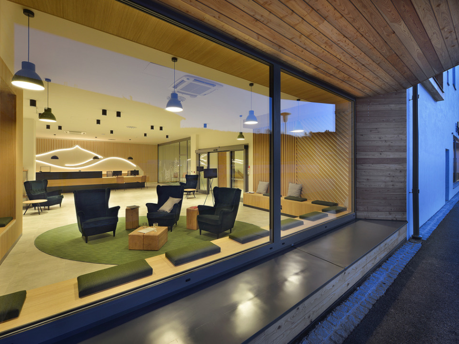 Modern glass-walled lobby with blue armchairs, wooden coffee tables, and a green circular rug in a contemporary reception area inching toward a bright, inviting space.