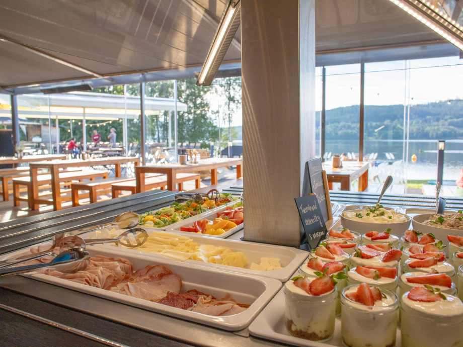 Buffet spread with sliced meats, cheeses, and fruit desserts, set up indoors with a lakeside view outside the windows.