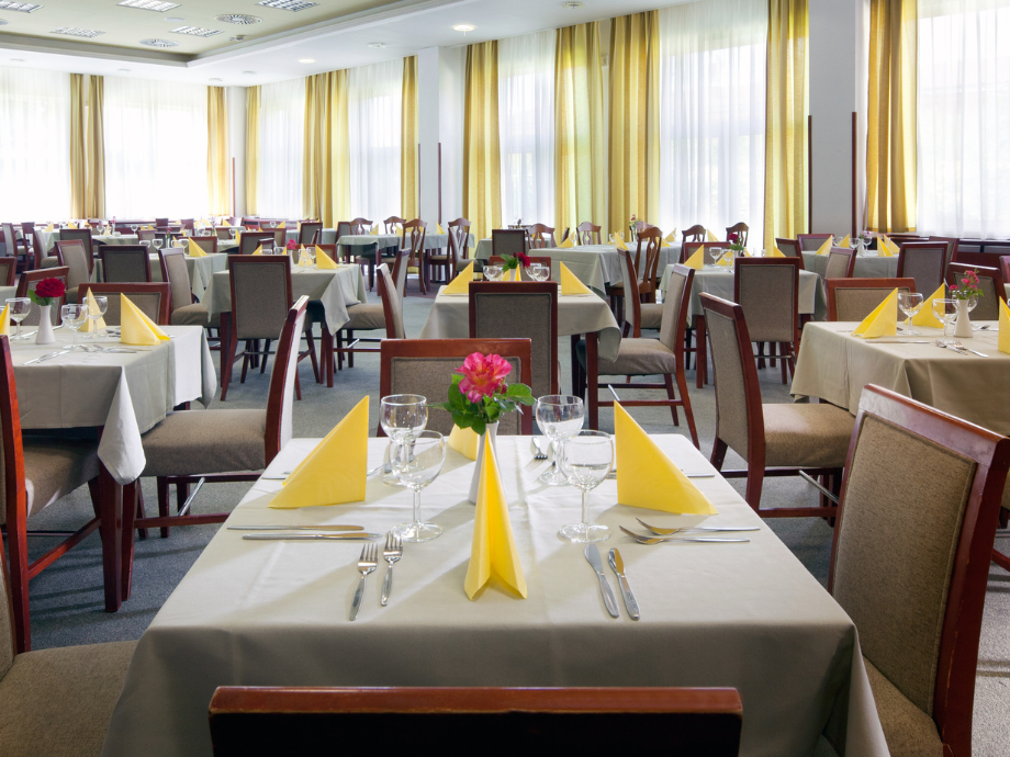 Banquet hall with many round and rectangular tables set for a meal, yellow napkins, glassware, and pink centerpieces under bright windows.