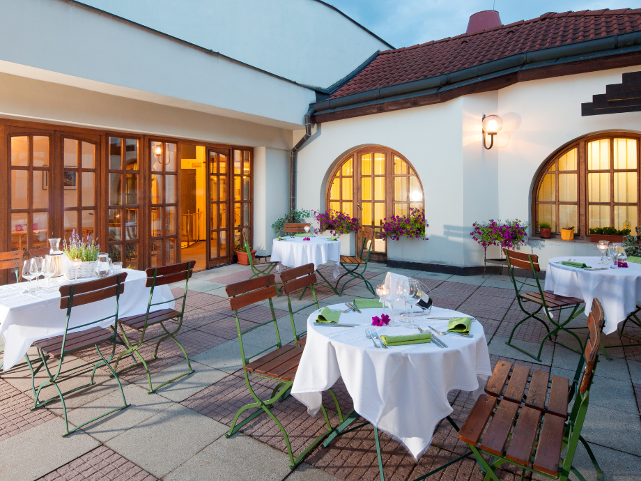 Outdoor cafe courtyard with white-draped tables, wooden chairs, and potted purple flowers against a white building with arched windows and warm interior lights.