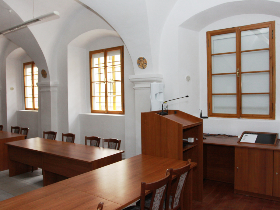 Lecture room with wooden podium, desk, and chairs, set against bright arched white walls and windows.",