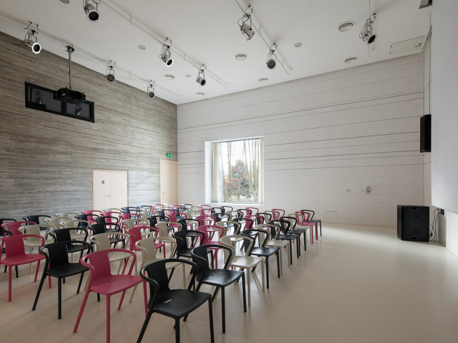 Empty lecture hall with rows of pink, beige, and black chairs, neutral walls, and a large back window.