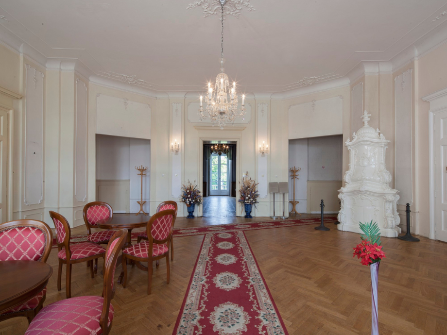 Formal reception room with a crystal chandelier, red-upholstered chairs around round tables, and a white ornate stove on the right.