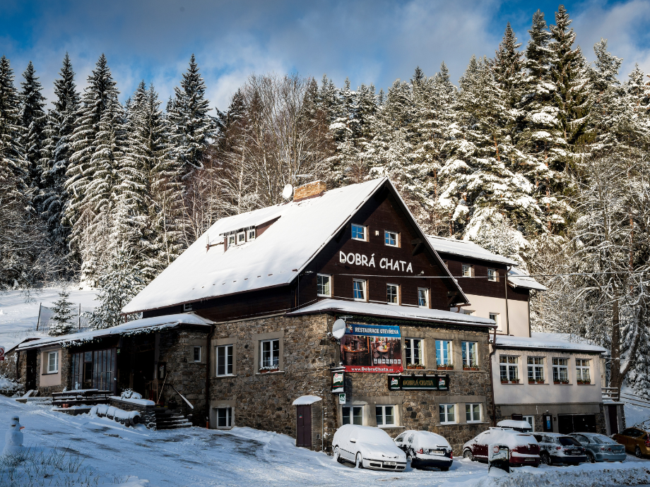 Snow-covered lodge named Dobrá Chata surrounded by snow-dusted pine trees in a forest.