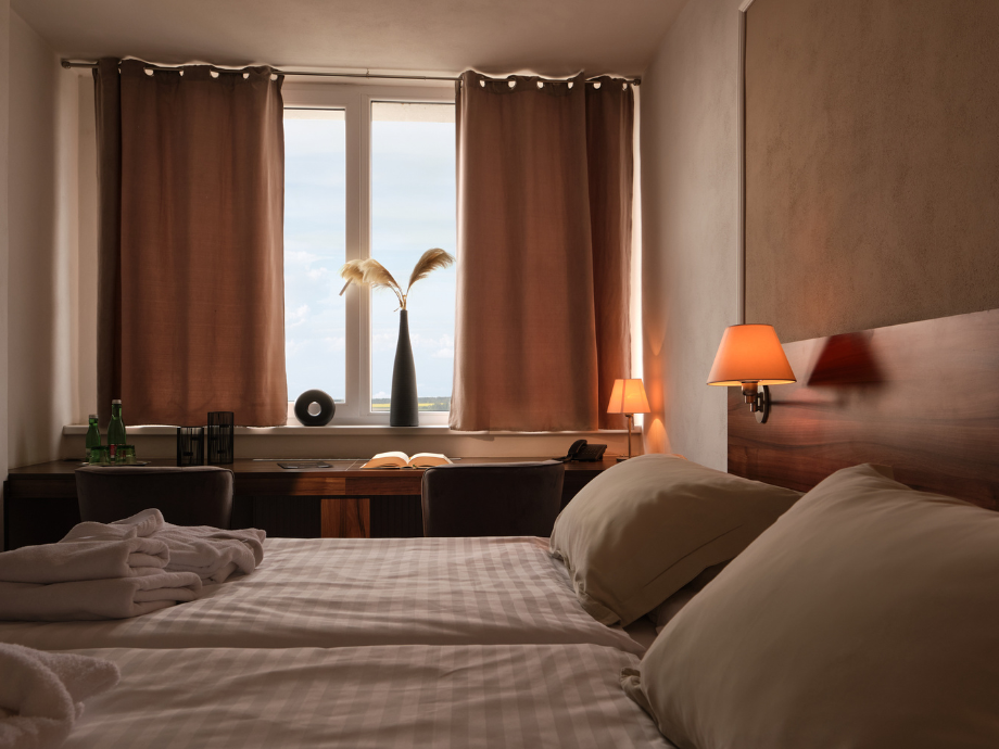 Hotel room featuring a made bed, two pillows, warm bedside lamps, and a desk by a window with brown curtains and a vase with tall feathers.