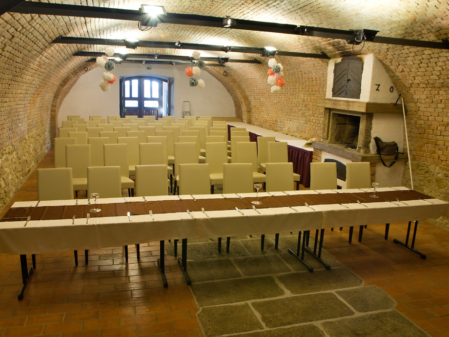Underground brick-vaulted room set for an event with rows of beige chairs and a long banquet table with glassware, facing a doorway at the back. The arched ceiling is decorated with hanging flowers, and a large brick fireplace sits to the right.