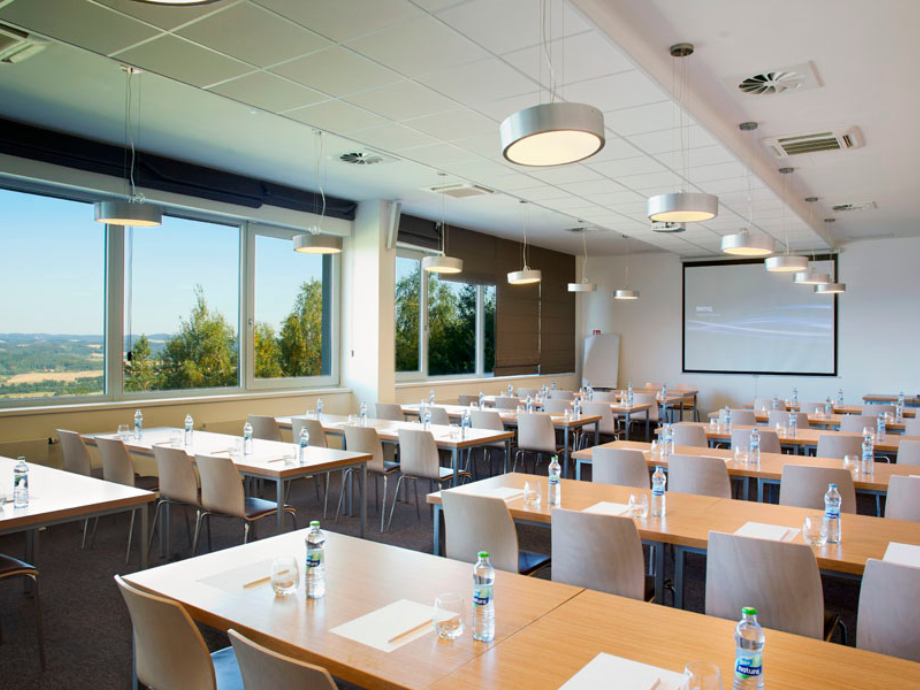 Bright conference room with rows of tables and chairs, water bottles and notepads, and a projector screen at the front.