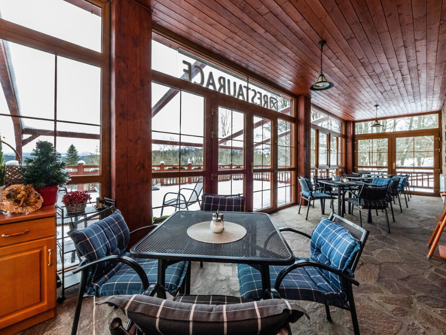 Covered outdoor seating area with blue plaid cushioned chairs around metal tables; snow outside the glass doors.