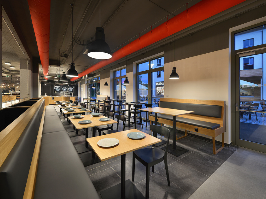 Modern dining area with wooden tables, black chairs, bench seating along a long counter, and red exposed ductwork under pendant lights.