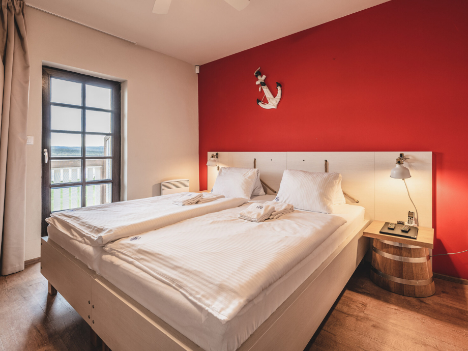 Hotel room with two single beds pushed together, white linens, red accent wall, anchor decor, and bedside lamps.