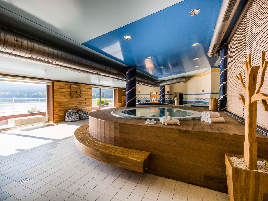 Indoor spa with a round wooden hot tub on a platform, towels folded nearby, and large windows overlooking water outside.