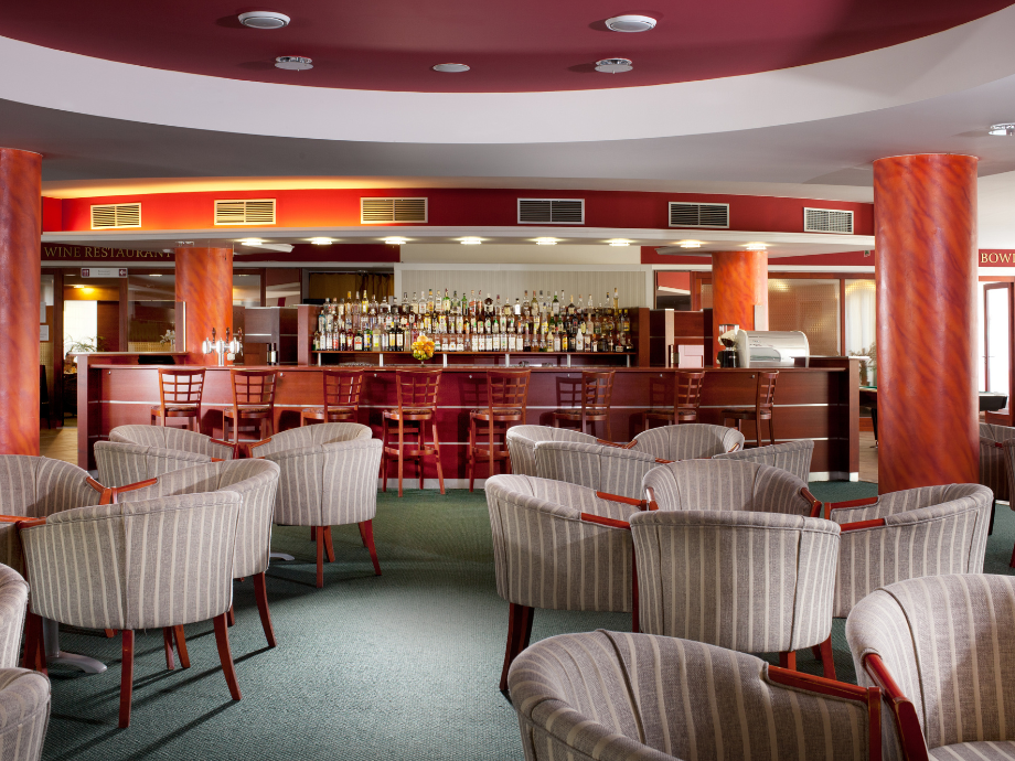 Hotel lounge bar with curved red ceiling, row of bottles behind a wooden bar and striped upholstered chairs before it of varying angles.