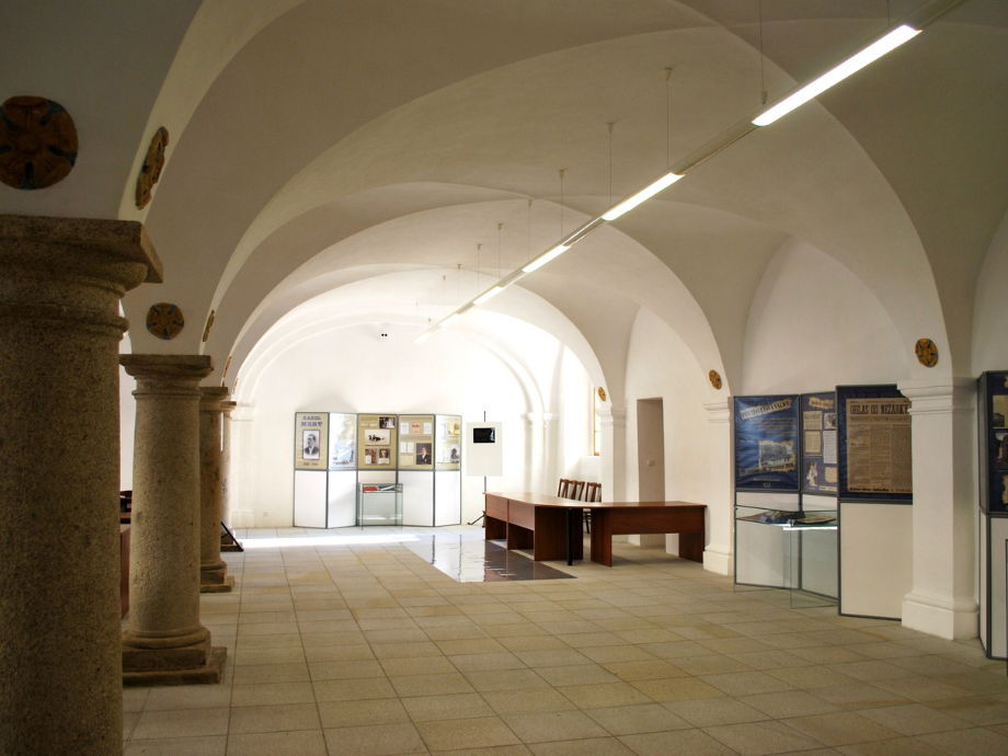 Museum hall with vaulted white arches, stone columns, and display boards along the back wall.