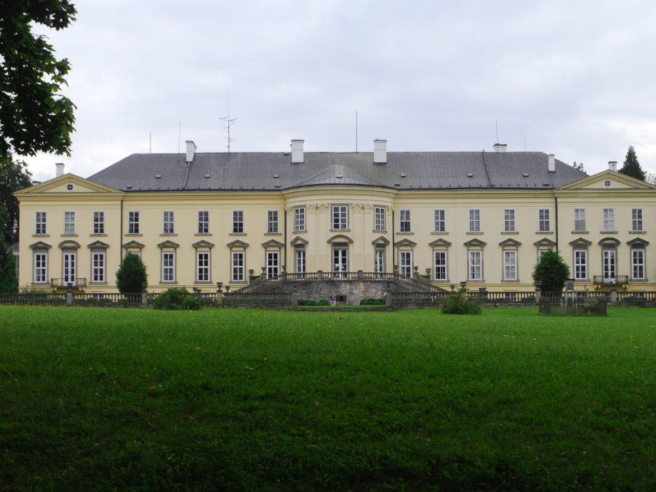 Front view of a large pale-yellow manor with a central curved staircase and symmetrical wings on a manicured lawn under a cloudy sky.