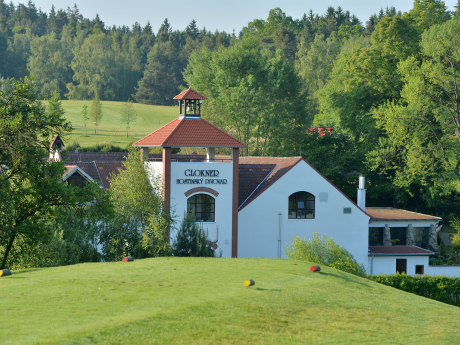 White brewery building with a red-tiled roof set in a green landscape, sign indicating a local brewery in the foreground.