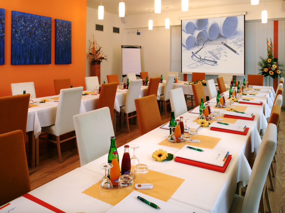Conference room arranged for a formal meeting or banquet with long white tables, orange and white chairs, and place settings ready for attendees.