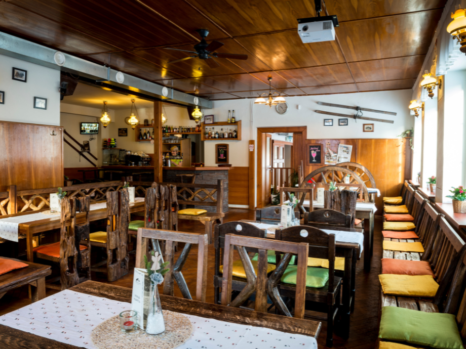 Cozy rustic restaurant interior with dark wooden tables and chairs, colorful cushion seating along a windowed bench, and a bar in the back.