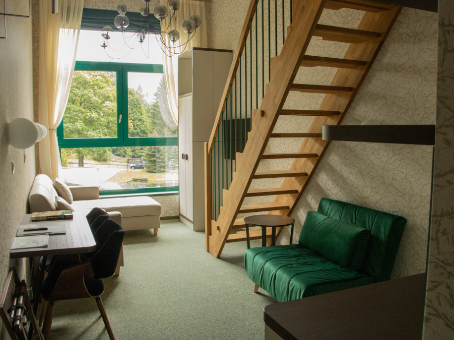Living room with a green-framed window letting in sunlight, a wooden staircase, and a green cushioned sofa facing a small round table.