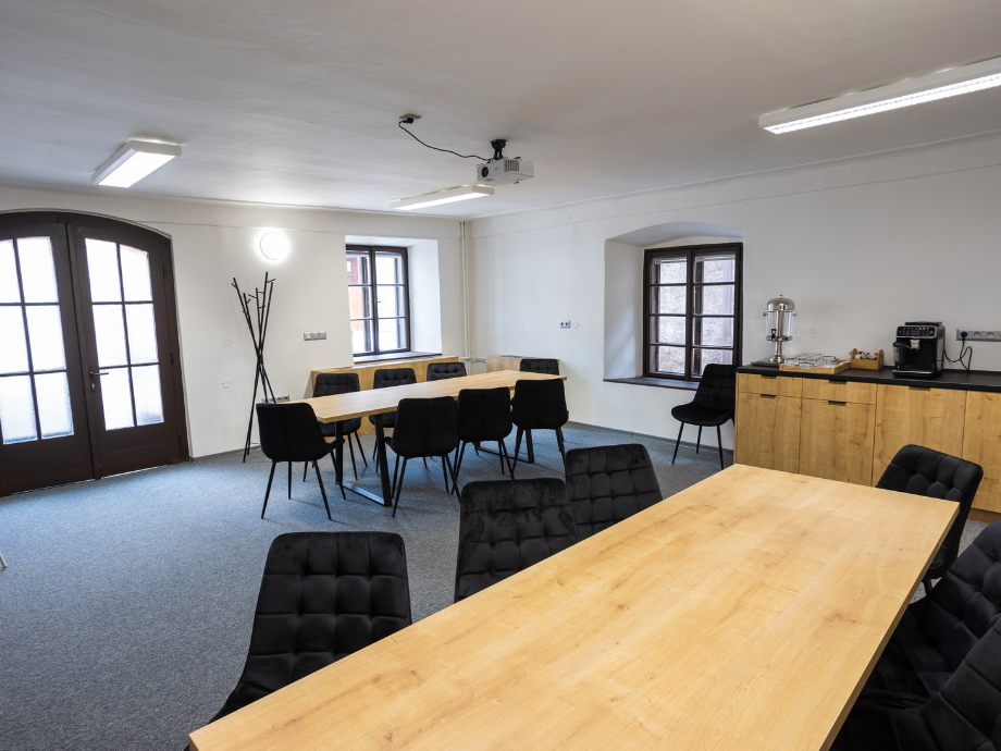 Conference room with two rectangular wooden tables and black upholstered chairs, projector on the ceiling, and a coffee station along the back wall.