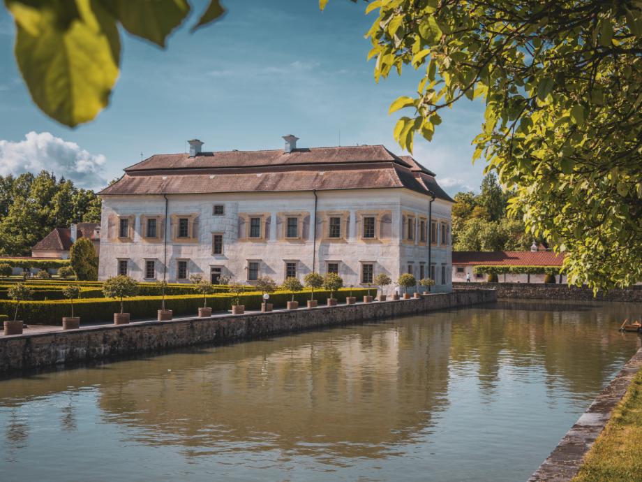 Historic white manor by a calm canal, with a manicured hedge garden and a red-tiled roof under a blue sky.
