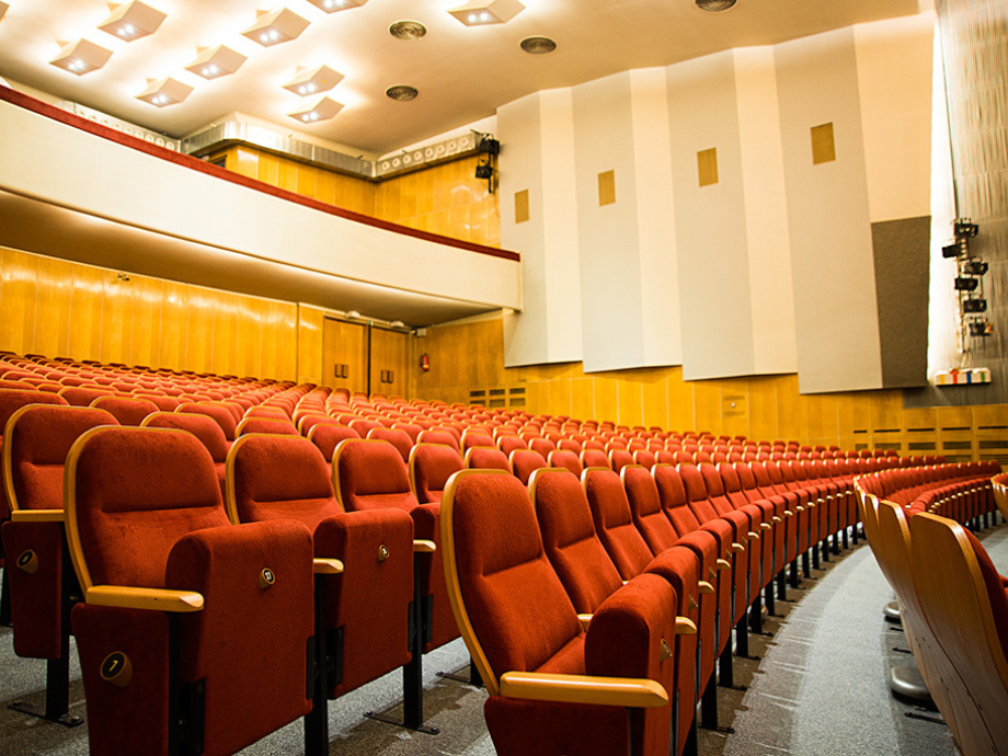 Empty theater auditorium with red upholstered seats and curved rows under warm ceiling lights.