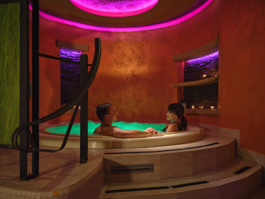 Couple relaxing in a circular hot tub in a dim spa with vibrant pink and purple lighting.