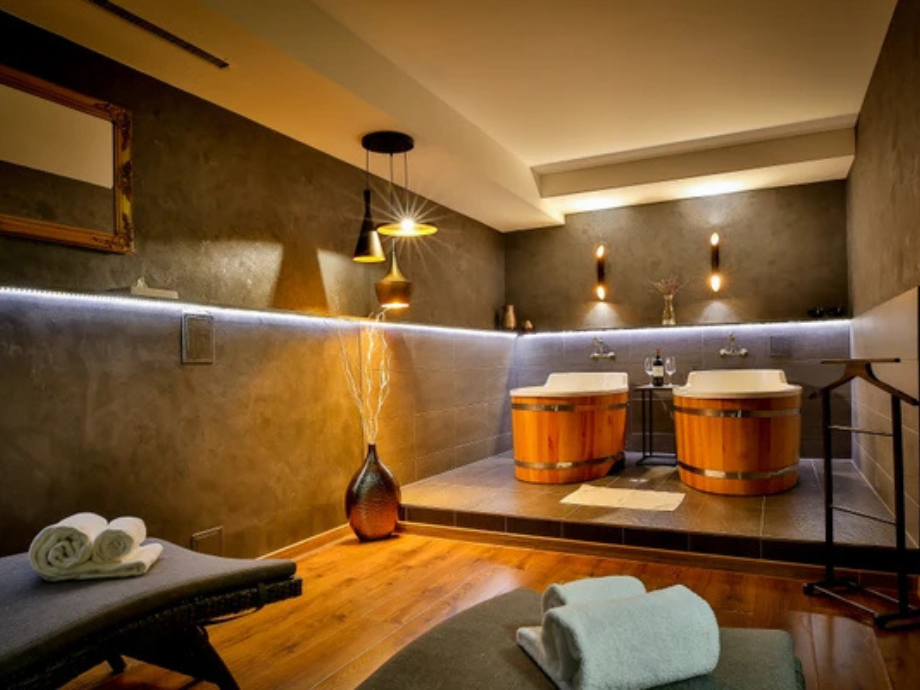 Spa room with two wooden soaking tubs on a tiled platform, warm ambient lighting, and rolled towels nearby