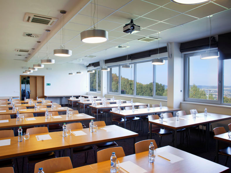 Bright conference room with long wooden tables, bottled water, and notepads, facing large windows with a tree-filled view.