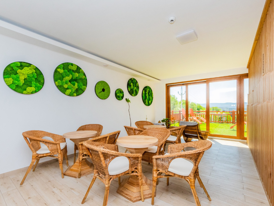 Bright indoor seating area with wicker chairs around round wooden tables, and circular green leaf wall art near glass doors to a sunny outdoor deck.