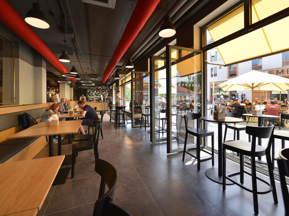 Cafe interior with wooden tables and black chairs; glass doors open to sunny outdoor seating under umbrellas.