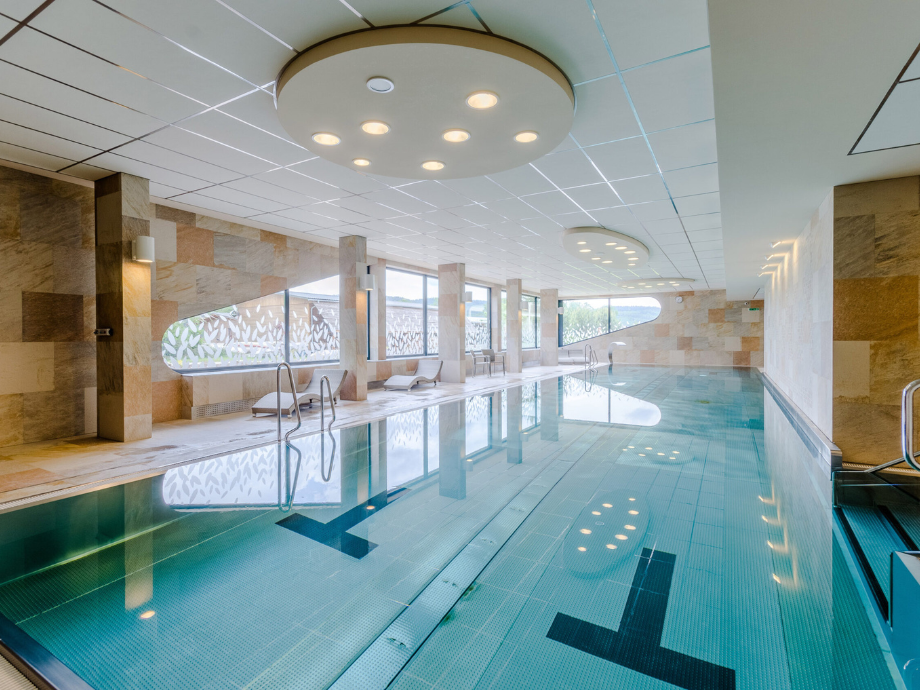 Indoor swimming pool in a spa-like area with round ceiling lights, beige stone walls, and lounge chairs along the pool edge.