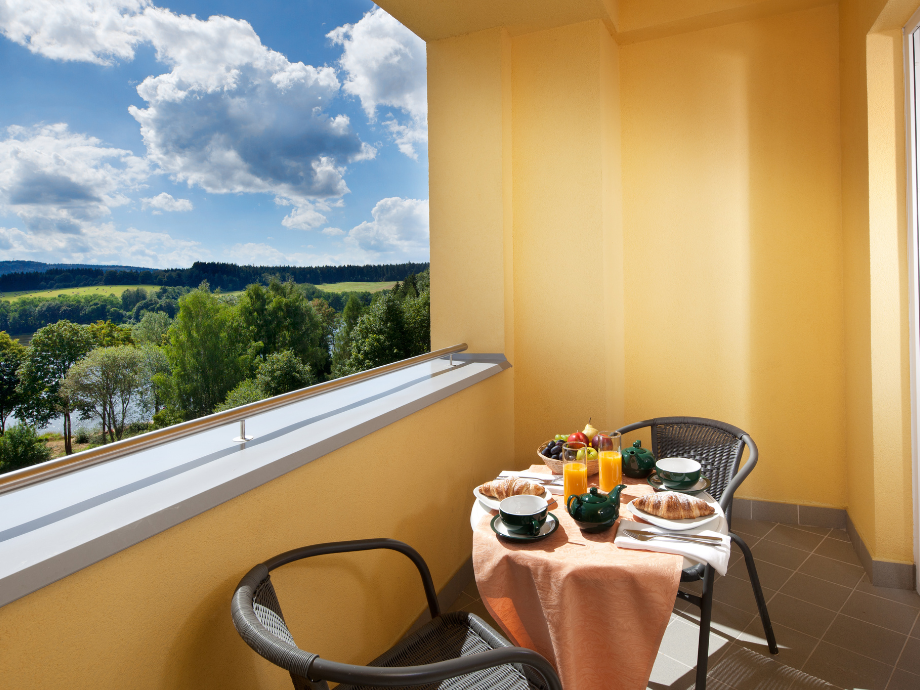 Sunny balcony with yellow walls set for breakfast: croissants, juice, tea cups, fruit, and a teapot on an orange tablecloth with two chairs, overlooking green landscape and a river.