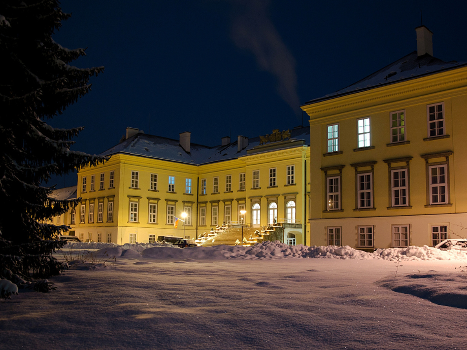 Nighttime view of a yellow historic building with snow-covered ground and lit steps leading to entrances, under a dark blue sky.