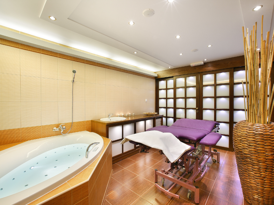 Spa treatment room with a large whirlpool tub and a padded massage bed, soft purple sheets, and warm ambient lighting.