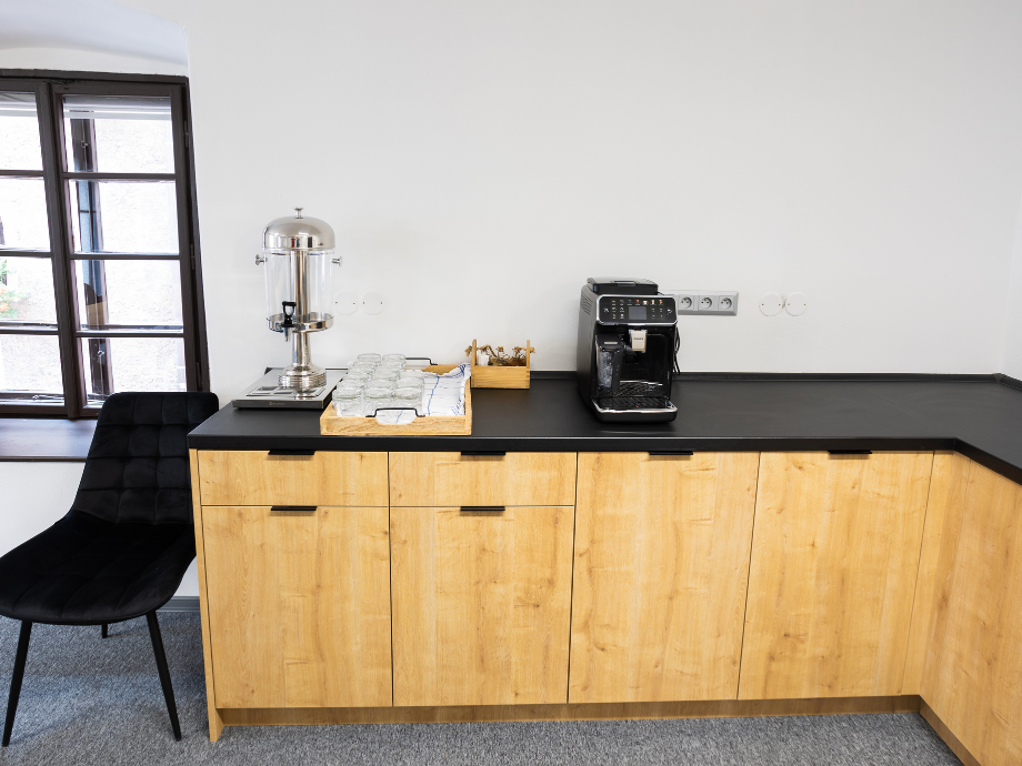 Coffee station with a stainless steel beverage urn, glass cups on a tray, and a black espresso machine on a dark countertop against a white wall.