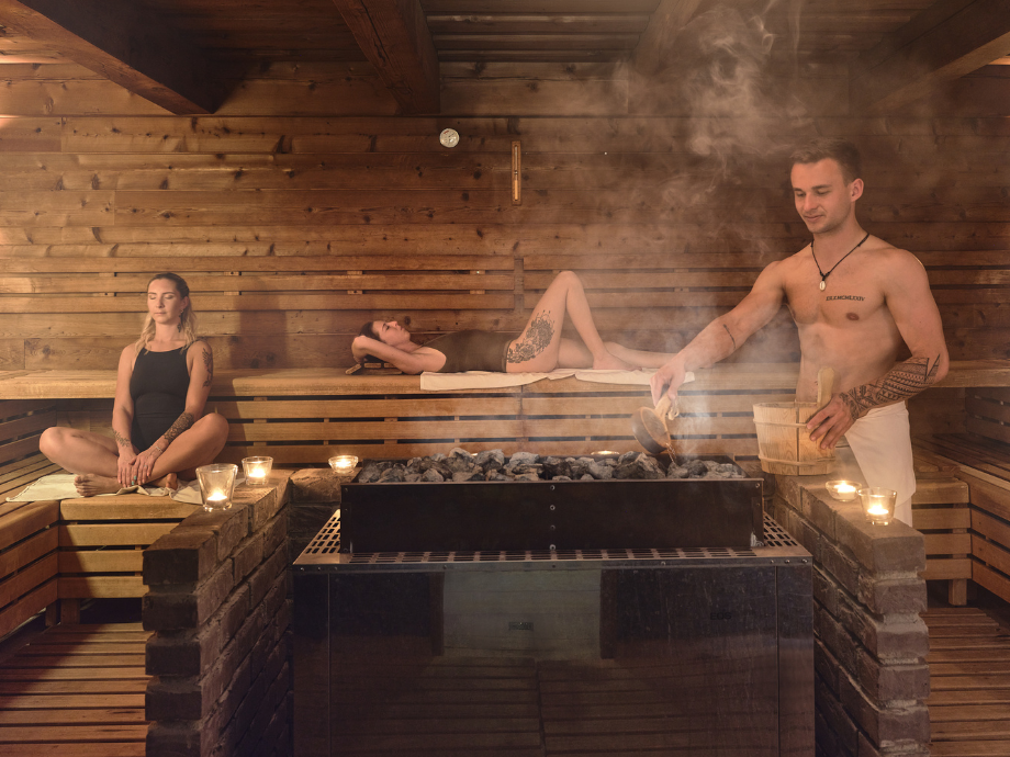 Three people in a wooden sauna; a woman sits cross‑legged left, a person rests on a bench center, and a shirtless man adds water to the rocks on the right.