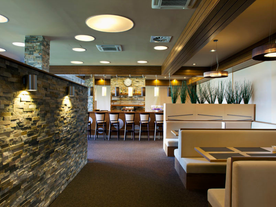Modern restaurant interior with a long bar, high stools, stone accent wall, and booth seating area.