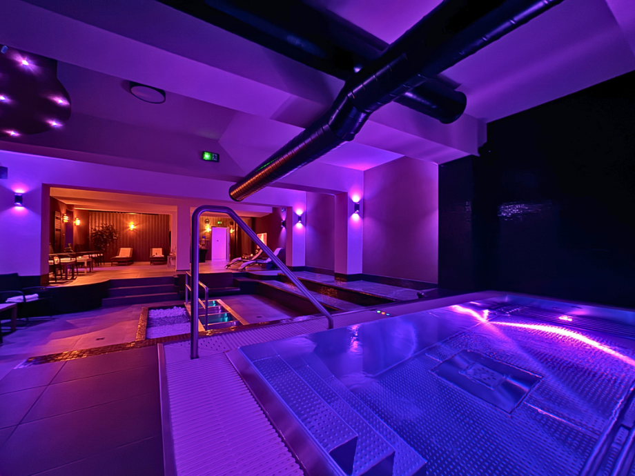 Indoor luxury spa with purple lighting, a small pool/hot tub, metal handrail, and a lounge area in the background.