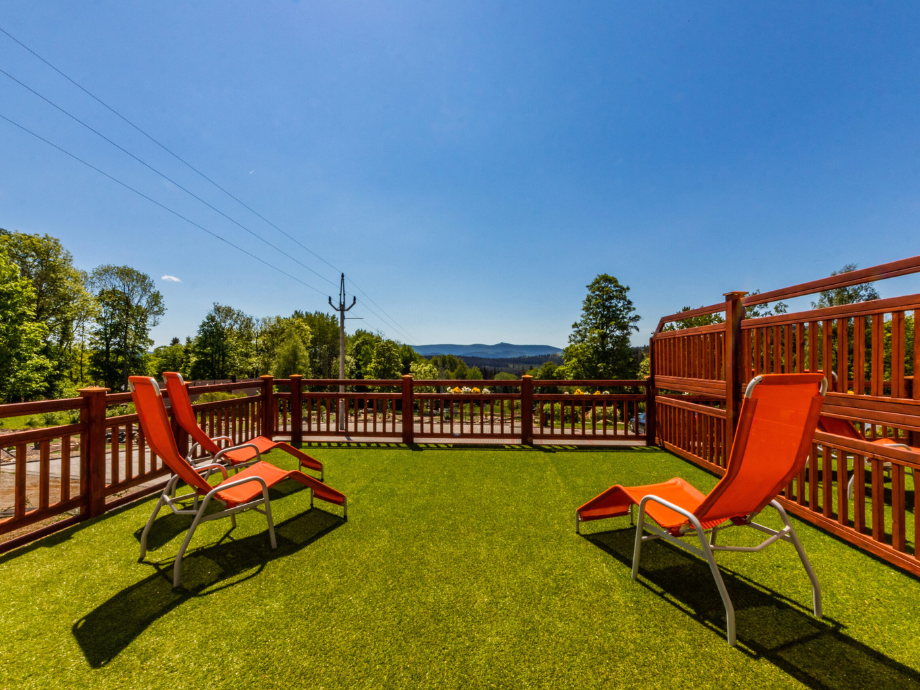 Sunny wooden deck with three orange lounge chairs on green grass, railing, and distant hills under a clear blue sky.