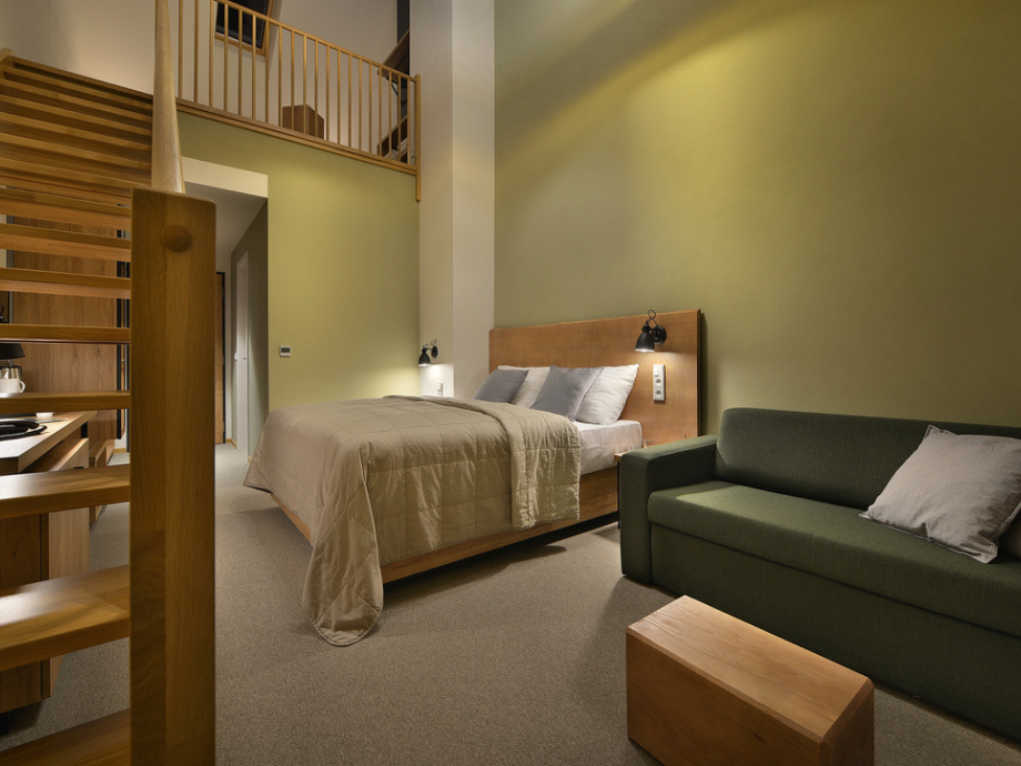 Hotel room with a wooden bed, beige bedding, green accent wall, and a green sofa in a lofted suite.
