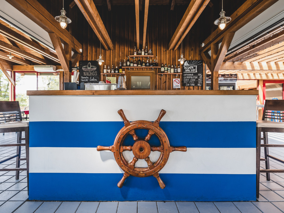 Bar counter with blue and white stripes and a wooden ship wheel, nautical decor in a wood-paneled room.