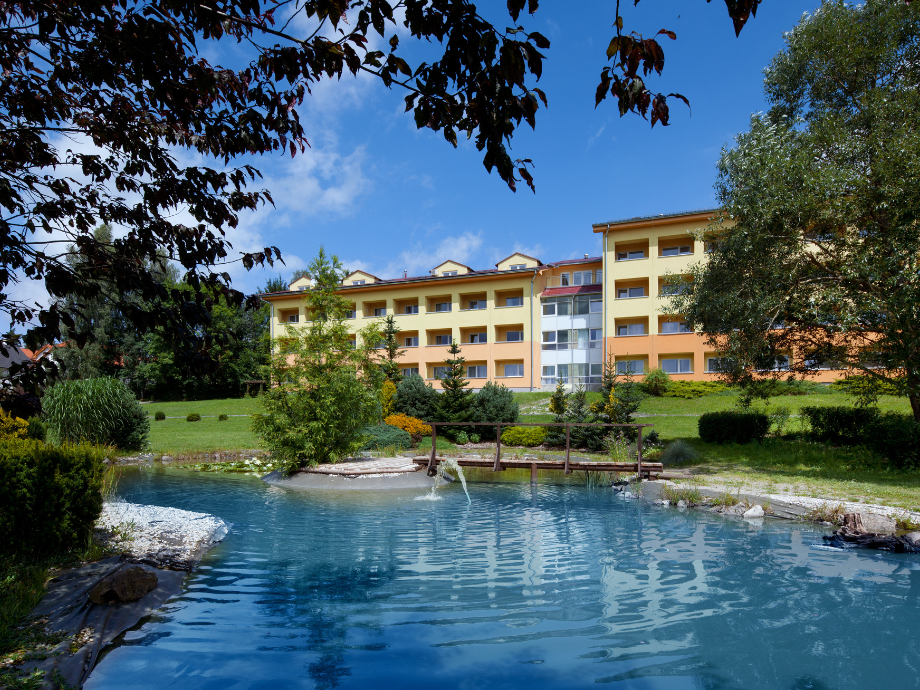 Yellow multi-story residential building with orange accents, flanked by a manicured garden and a small wooden bridge over a blue pond in bright daylight.