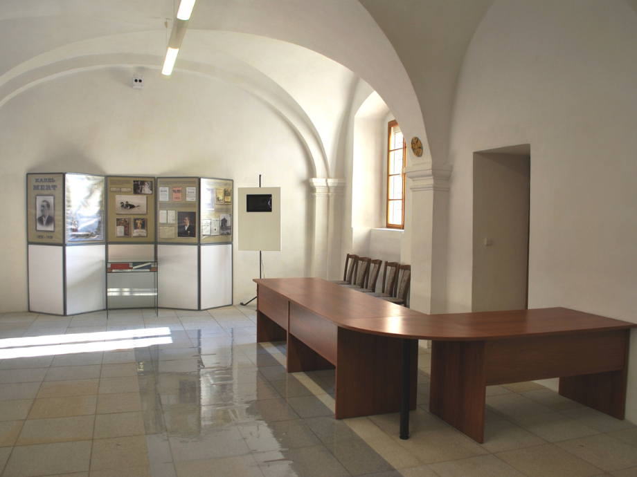 Museum exhibit room with display panels, a long curved wooden desk, and a row of chairs under a vaulted ceiling
