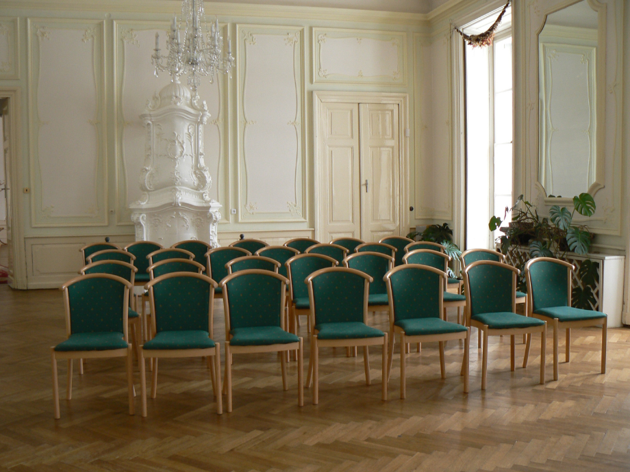 Elegant historic room with mint-green walls, a white ornate heater, and rows of green upholstered chairs set up for an event.